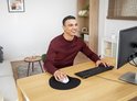 Tapis de souris noir avec repose-poignet, avec une souris dessus, sur un bureau en bois, pendant qu'un homme travaille sur un ordinateur en souriant.