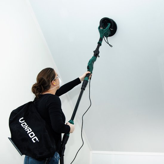 Vrouw met rugzak schuurt een hoog plafond met een lange groene schuurmachine.