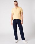Man in een geel poloshirt en donkerblauwe spijkerbroek, staand.