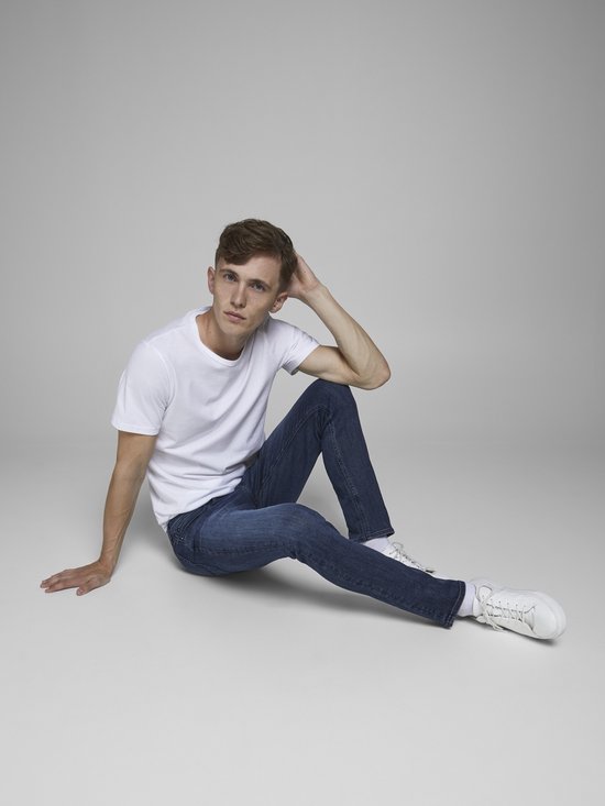 Man in een wit t-shirt en donkerblauwe jeans, zittend op de grond.