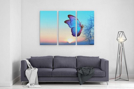 Drieluik schilderij met een blauwe vlinder op een paardenbloem tegen een zonsondergang achtergrond, boven een grijze sofa.
