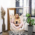 Vrouw in lichte roze outfit zit ontspannen in een witte hangstoel met macramé en franjes.