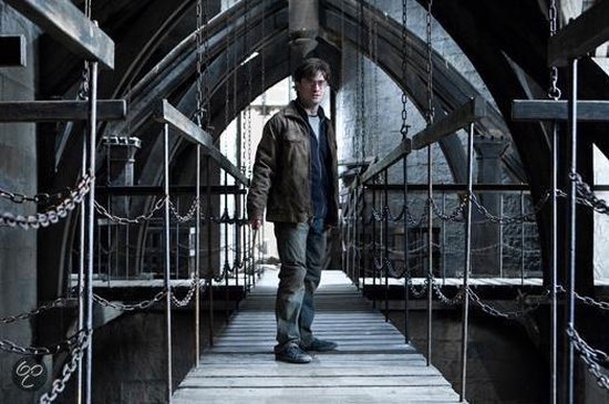 Harry Potter, met een bruine jas, staat op een houten brug met kettingen, in een donkere, architectonische omgeving.