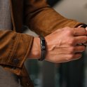 Close-up van een man met een bruine gevlochten leren armband met witte stiksels en zwarte sluiting met leeuwenlogo.