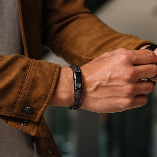 Close-up van een man met een bruine gevlochten leren armband met witte stiksels en zwarte sluiting met leeuwenlogo.