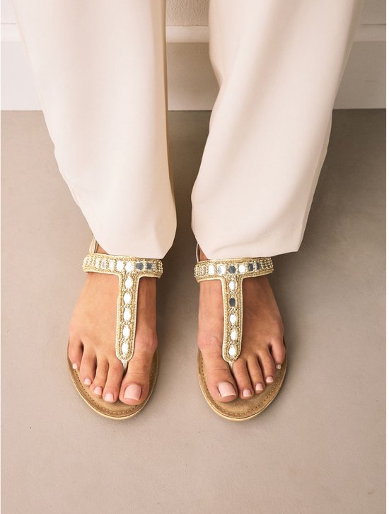 Voeten in gouden sandalen met platte zool, versierd met parels en spiegeltjes op de bandjes en teenstuk.