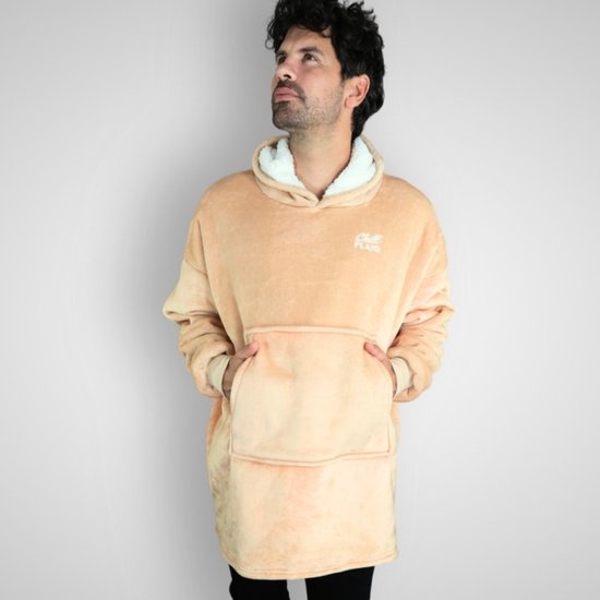 Een man met krullend bruin haar staat met zijn handen in de zakken van een beige hoodie deken. Hij kijkt omhoog.