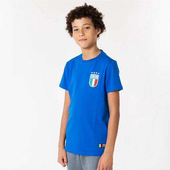 Jongen met krullend haar in een blauw T-shirt met "ITALIA" embleem, staand met handen in de zakken.
