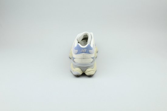 Achterkant van een lichtblauwe en beige New Balance sneaker, staand op de grond.