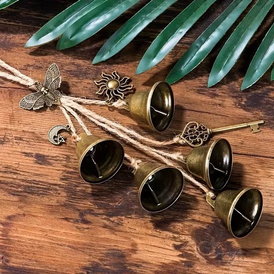 Bronzen heksenbel met belletjes, vlinder, sleutel, zon en maan, liggend op een houten ondergrond met groene bladeren.
