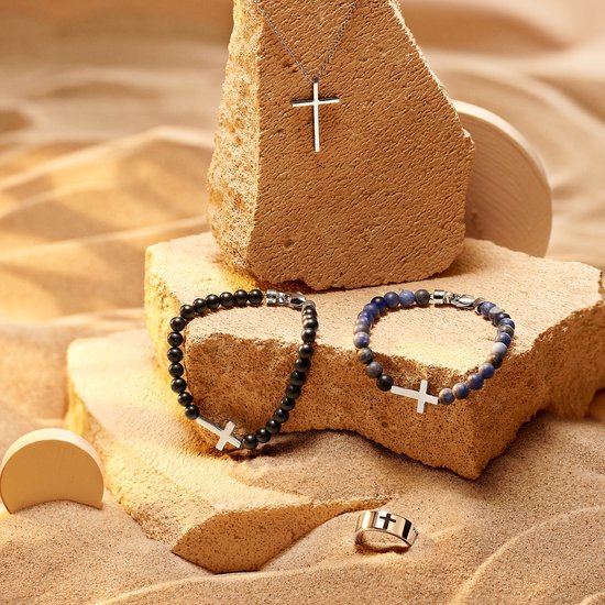 Zilverkleurig kruis ketting, armbanden met zwarte en blauwe kralen, en een ring op zand.