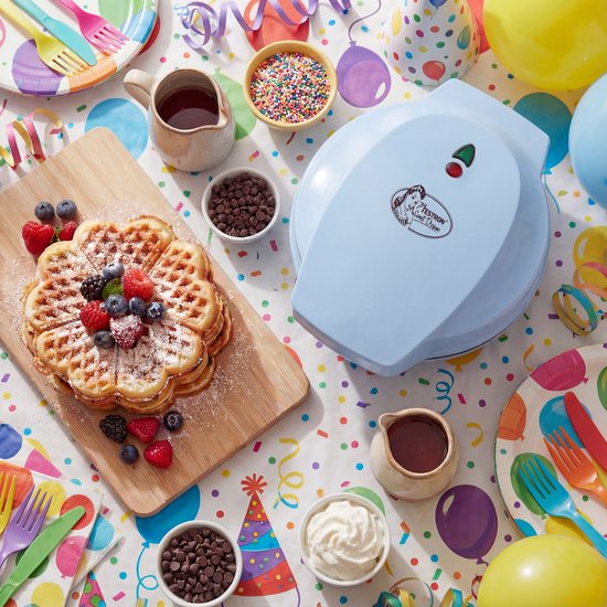 Blauw wafelijzer op feestelijk gedekte tafel met grote hartvormige wafel, fruit, toppings en ballonnen.