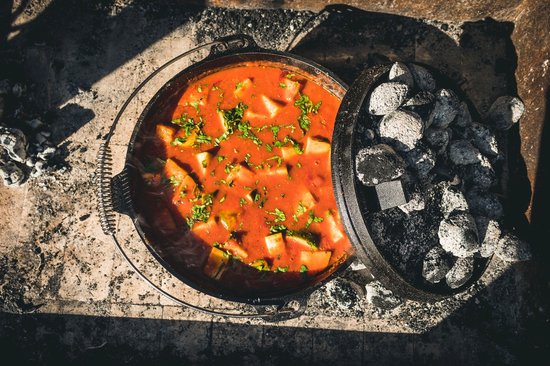 Gietijzeren pan met rode stoofpot, gegarneerd met kruiden, met kolen op het deksel.