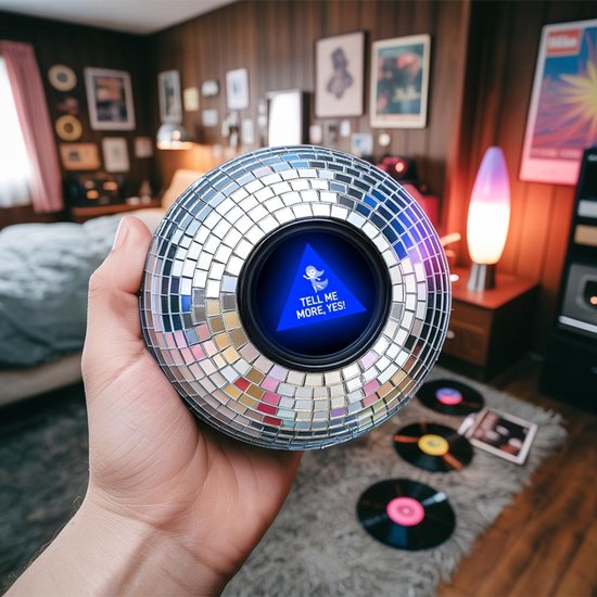 Hand die een discobal vasthoudt met een scherm dat 'TELL ME MORE, YES!' zegt. Op de achtergrond staat een slaapkamer.