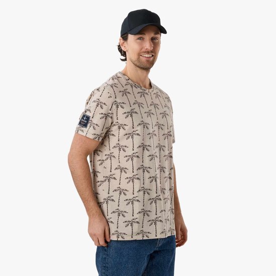 Man glimlacht en draagt een beige T-shirt met palmbomenprint, spijkerbroek en zwarte pet.