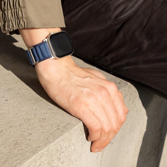 Donkerblauw nylon bandje om een pols met een goudkleurige Apple Watch. De pols is bij een grijze betonnen rand.