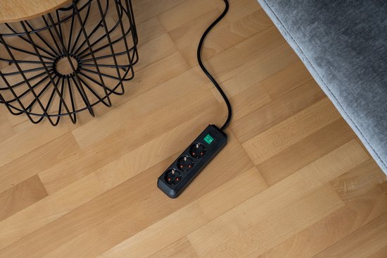 Zwarte stekkerdoos met groene schakelaar op een houten vloer, naast een bijzettafel en bank.