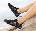 Zwarte waterschoenen met zilverkleurige lijnen en witte veters, gedragen door iemand op een rotsachtig strand.