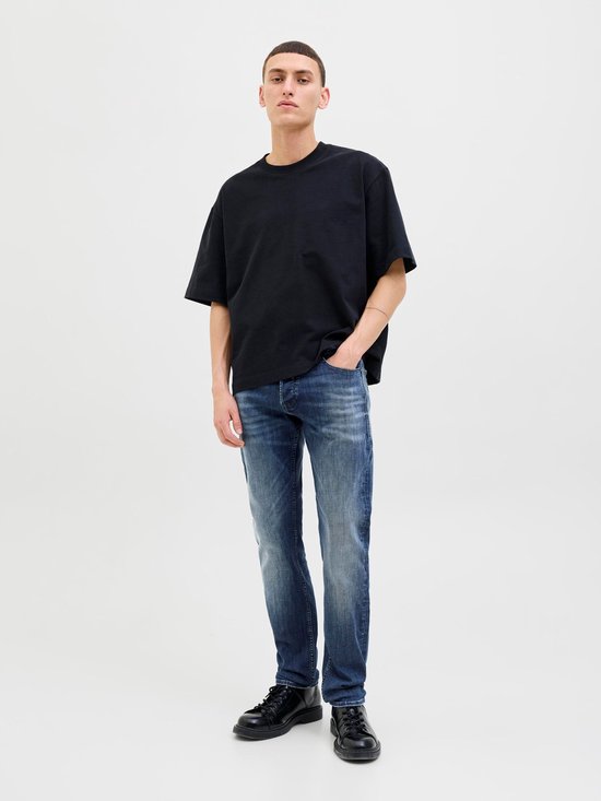 Man in een zwart oversized T-shirt, blauwe jeans en zwarte schoenen, met de hand in de zak.