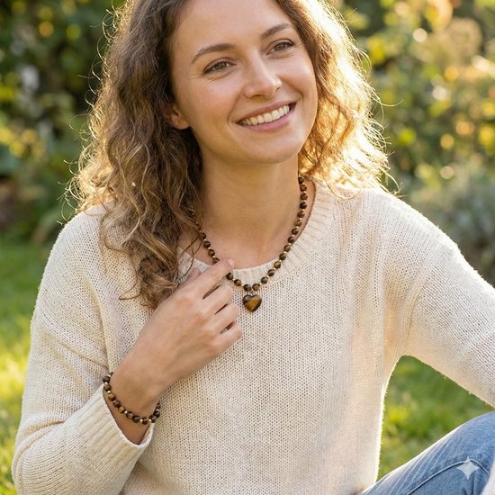 Vrolijke vrouw draagt een bruin-zwarte kralenketting met hartvormige tijgeroog hanger en gouden accenten.