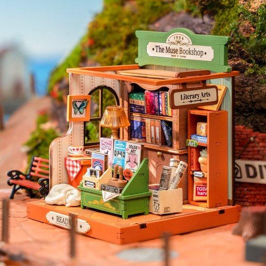 Miniatuur boekenwinkel model, "The Muse Bookshop", met boeken, lamp, kat en bankje, in een buitenomgeving.