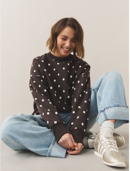 Model zit op de grond in een lichte jeans en donkerbruine sweater met witte polkadotprint.