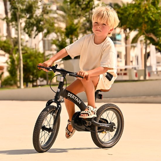 Petit garçon aux cheveux blonds sur un vélo Deryan noir 3-en-1 avec guidon et selle marrons, assis sur une place ensoleillée.