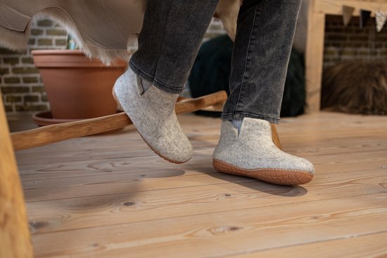 Persoon met grijze jeans draagt lichtgrijze vilten pantoffels met bruine zolen op een houten vloer.