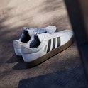 Paar witte Adidas sneakers met zwarte strepen en bruine zolen, op een betonnen ondergrond.