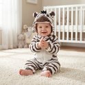 Baby in zebra onesie zit op een vloerkleed bij een ledikant.