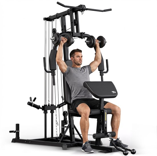 Man in grijs T-shirt en korte broek doet schouderoefening met dumbbells op zwart krachtstation.
