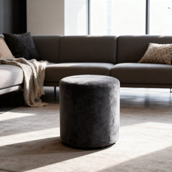 Donkergrijze fluwelen poef, rond, in een woonkamer met bank en kussens.