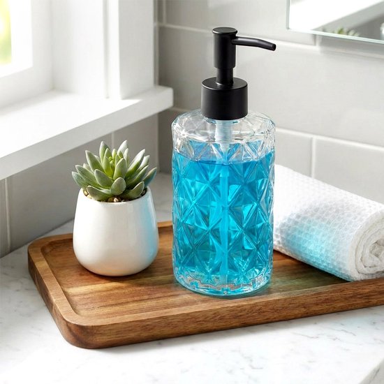 Glazen zeepdispenser met zwarte pomp, gevuld met blauwe zeep, naast een plantje en handdoek.