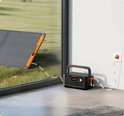 Donkergrijs Jackery Explorer 500 powerstation met oranje accenten, aangesloten op een stopcontact, met een zonnepaneel ernaast.