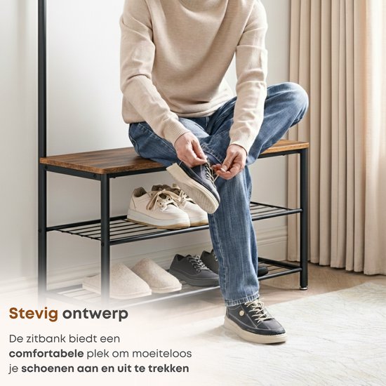 Man met spijkerbroek en beige trui zit op een bruine bank en doet schoenen aan. Schoenenrek met schoenen eronder.