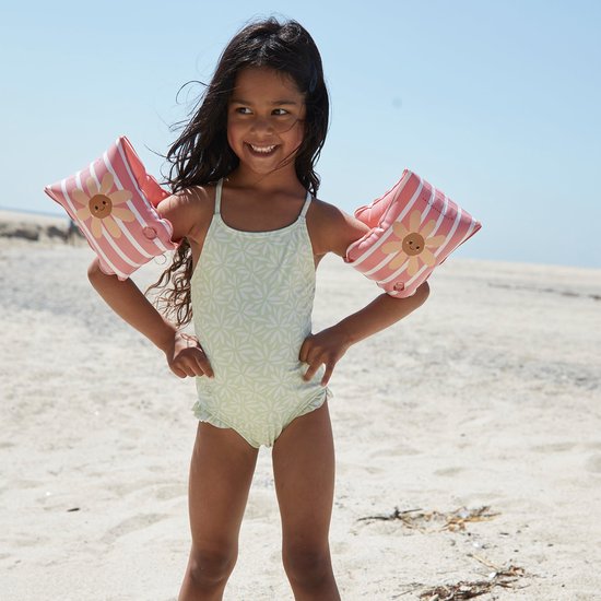 Kind op het strand met lichtgroen zwempak met bloemenprint en roze gestreepte zwembandjes met bloemmotief.