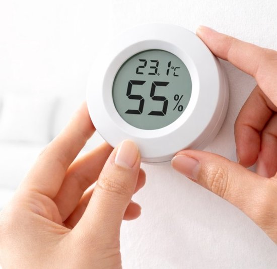 Witte digitale hygrometer/thermometer wordt door handen op een witte muur geplaatst, toont 23.1°C en 55%.