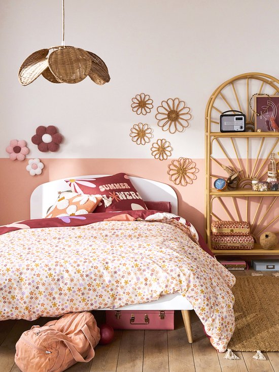 Une chambre d'enfant avec un lit, une parure de lit à imprimé floral, des fleurs murales en rotin, une étagère en rotin et une lampe en rotin.