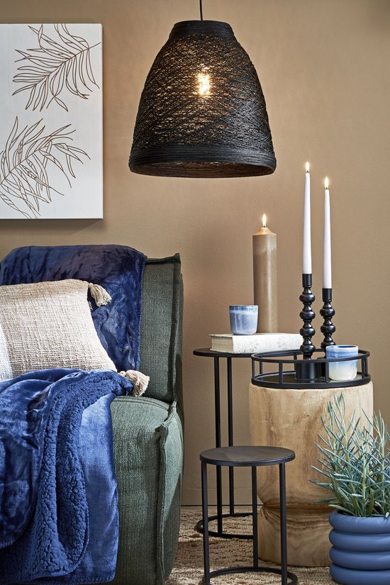 Zwarte rotan hanglamp boven een groene bank met een blauwe plaid en kussens.