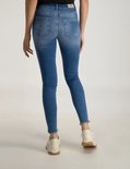 Achterkant van blauwe skinny jeans met lussen en rafelzomen, gedragen met witte sneakers.
