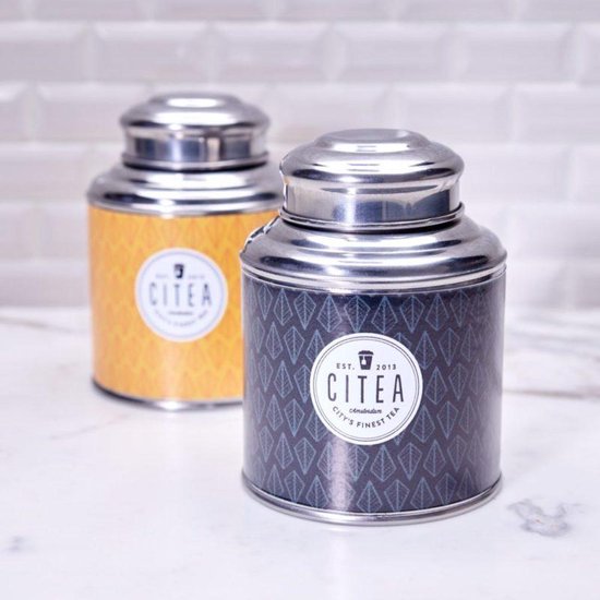 Twee ronde metalen theeblikken met CITEA logo. Eén blik is geel met ruitjespatroon, de ander donkerblauw met bladerenpatroon.