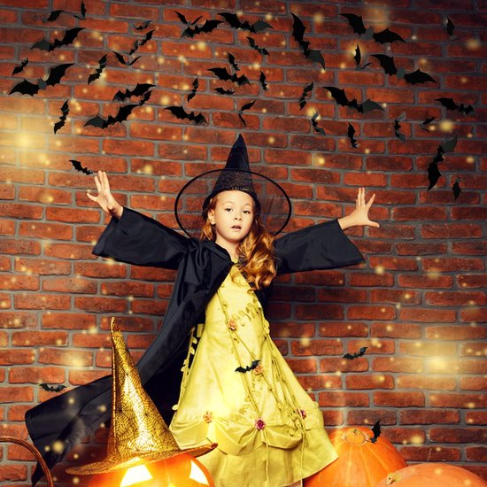 Une jeune fille en costume de sorcière avec un chapeau pointu se tient devant un mur de briques, avec des citrouilles et des autocollants chauve-souris.