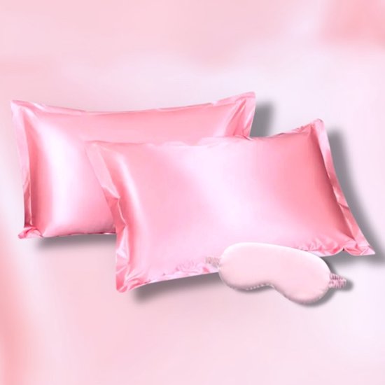 Twee lichtroze satijnen kussenslopen en een bijpassend slaapmasker op een roze achtergrond.