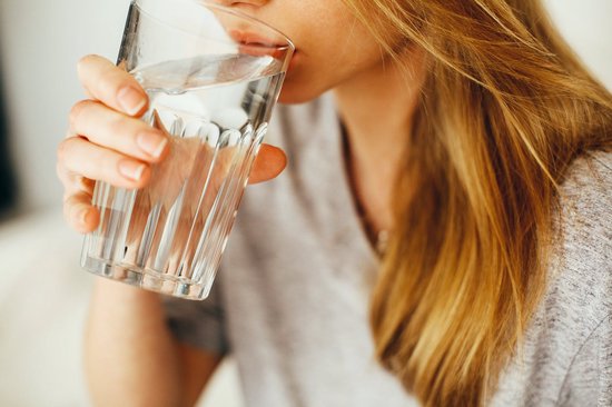 Een vrouw met blond haar drinkt uit een glas met water.