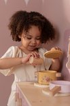 Kind met krullend haar in creme kleding speelt met roze speelgoedkeuken, houdt geel pannetje vast.