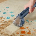 Donkerblauwe hand van een tapijtreiniger die een beige tapijt schoonmaakt met blauwe pootafdrukken en oranje vlekken.