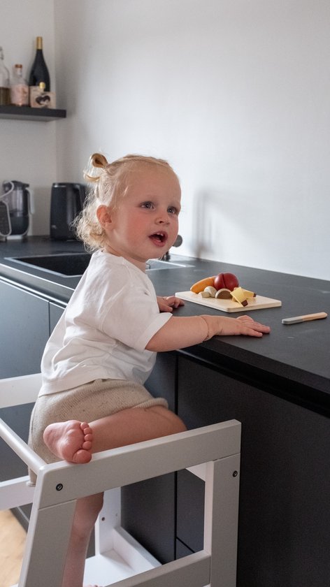 Kind met blonde knotjes in wit shirt en beige broek zit op witte leertoren, kijkt naar keukenblad.