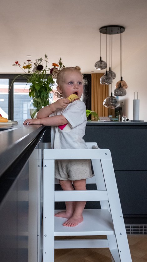 Kind met blonde knotjes in wit shirt en beige broek eet koekje op witte leertoren in keuken.