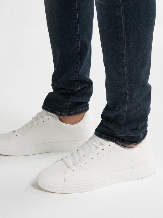 Close-up van de broekspijpen van een donkerblauwe jeans met witte sneakers.