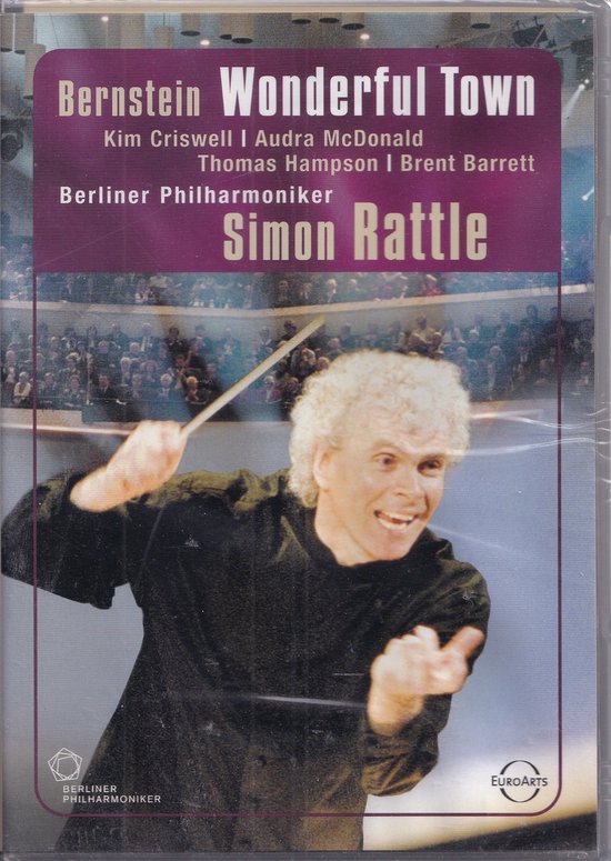 Dirigent Simon Rattle met warrig haar, zwarte kleding, gebaren met stok, orkest en publiek op achtergrond.
